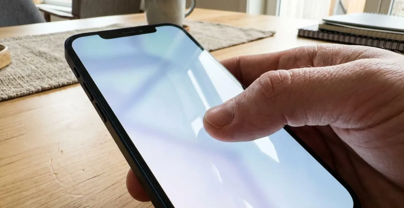 Primo piano estremo di una mano che digita su uno smartphone moderno, con lo schermo completamente sfocato e riflessi luminosi, su una superficie di legno chiaro pulita