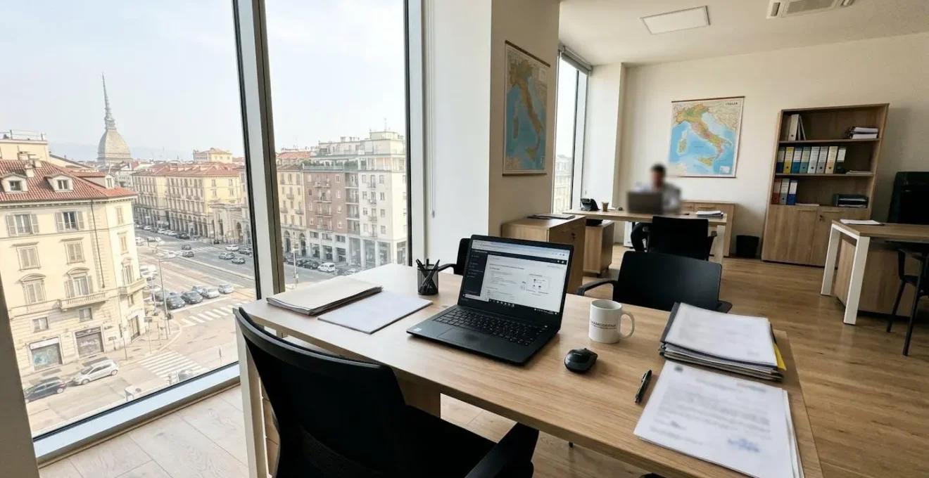 Interno di un ufficio immobiliare moderno italiano con scrivania ordinata, monitor e documenti parzialmente visibili ma sfocati, luce naturale dalla finestra e città italiana sullo sfondo
